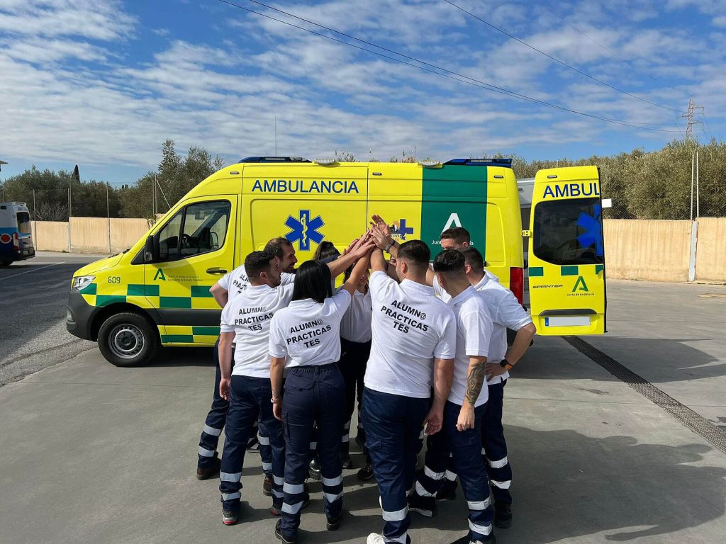 Alumnos de CETES de Técnico en Emergencias Sanitarias con el uniforme de prácticas uniendo sus manos frente a una ambulancia en la nave de ambulancias